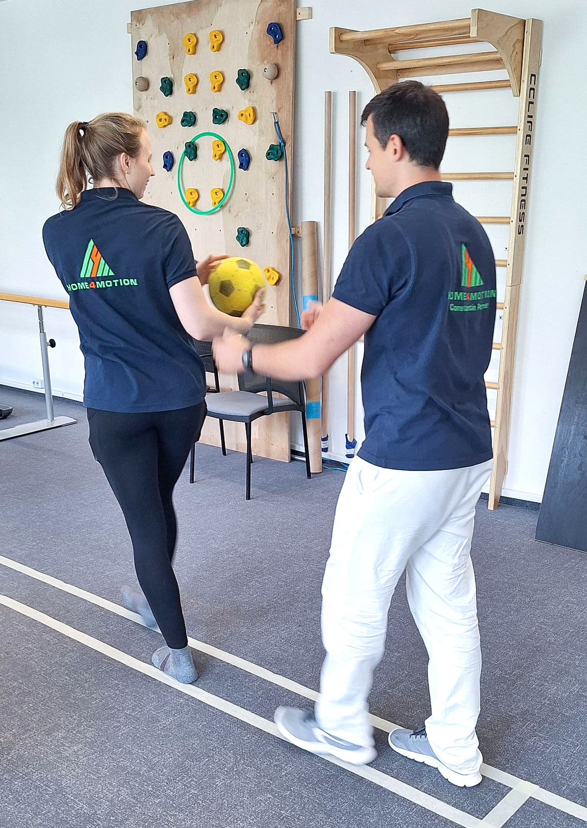im Hintergrund Kletterwand und Sprossenwand. Davor 2 Therapeut:innen die sich auf einer Linie einen Ball nach hinten geben